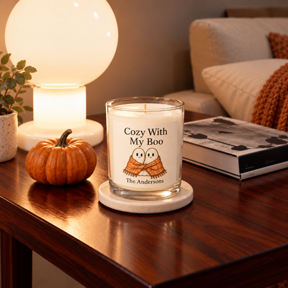 A Cozy With My Boo candle showing two friendly ghosts sharing an orange blanket and The Andersons as sample personalization, on a stone coaster beside a pumpkin, books, and a warm lamp.