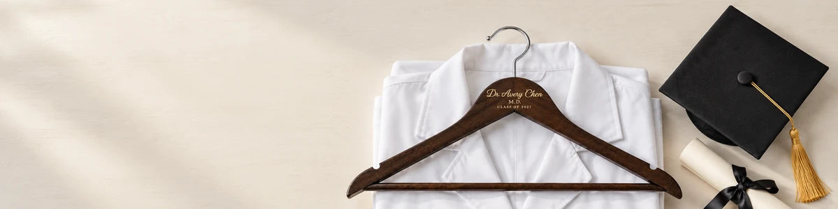 Personalized dark wood Dr. Avery Chen hanger resting on a folded white medical coat beside a black graduation cap and rolled diploma on a pale tabletop
