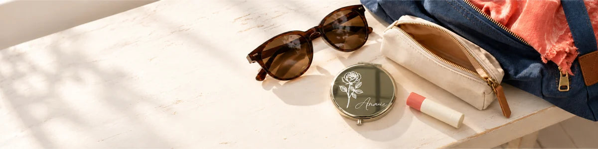 Personalized gold floral compact mirror with sunglasses, lip balm and a weekend bag on a pale bench